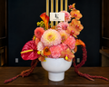 Sunset Bloom arrangement by Lenorah & Co. — dinner-plate dahlias, ruffled carnations, coral anthurium and burnt-orange calla lilies with trailing amaranth in a matte white vessel.