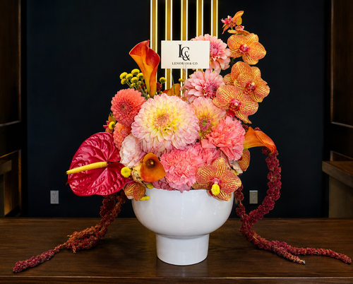 Sunset Bloom arrangement by Lenorah & Co. — dinner-plate dahlias, ruffled carnations, coral anthurium and burnt-orange calla lilies with trailing amaranth in a matte white vessel.